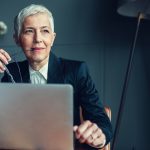 Mature Businesswoman In Her Office. Portrait of mature businesswoman, sitting in her office and thinking. Holding eyeglasses in her mouth. In front of her on the desk is laptop.