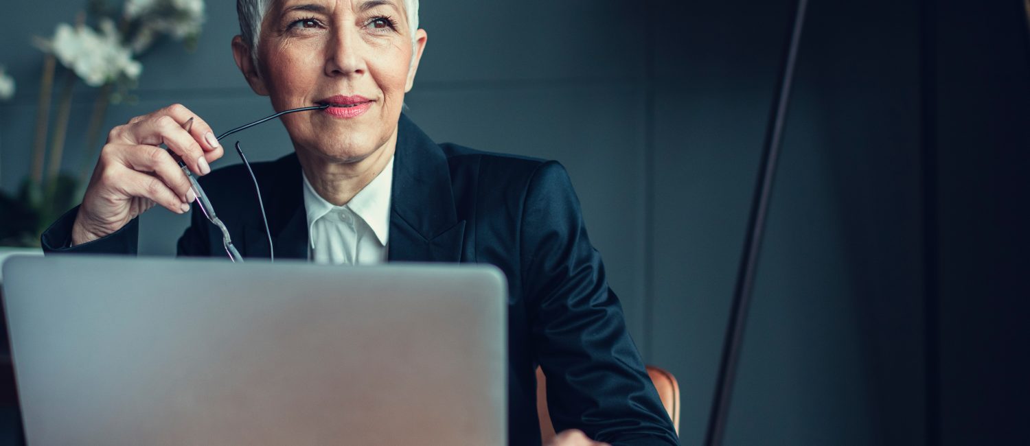 Mature Businesswoman In Her Office. Portrait of mature businesswoman, sitting in her office and thinking. Holding eyeglasses in her mouth. In front of her on the desk is laptop.